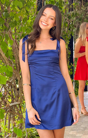 Model smiling in a navy blue satin mini dress, perfect for homecoming. The dress features a straight, pleated neckline and wide shoulder straps that tie into bows.