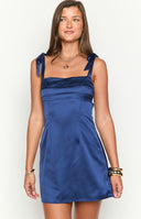 Model smiling in a navy blue satin mini dress, perfect for homecoming. The dress features a straight, pleated neckline and wide shoulder straps that tie into bows.
