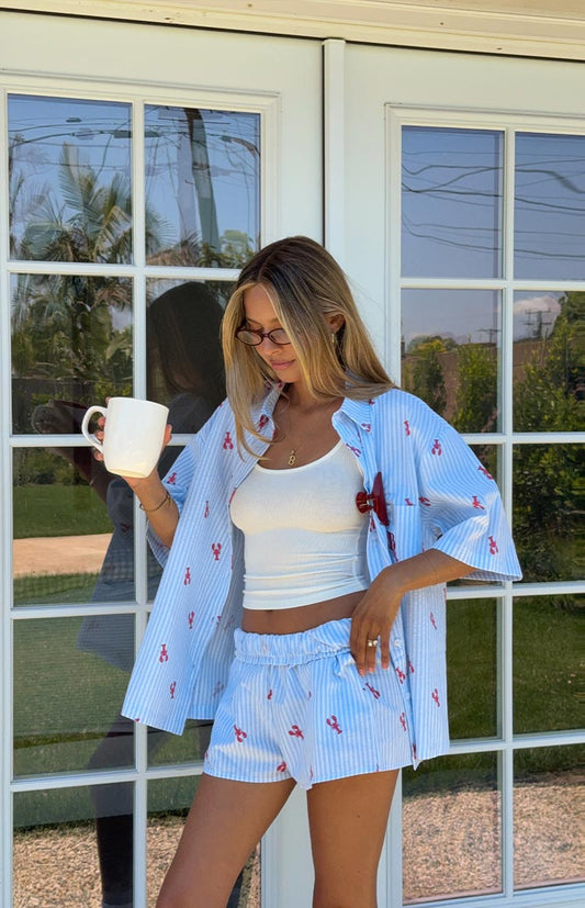 A woman wears a light blue and white striped pajama shirt with a red lobster print, unbuttoned over a white ribbed tank top, paired with matching blue and white striped shorts with the same lobster print.