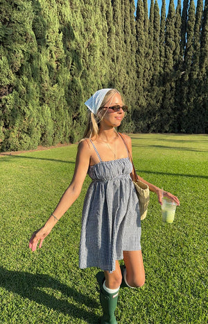 A woman in a black and white gingham mini dress with a ruched bust and spaghetti straps, a white bandana tied around her head, and dark green rain boots with white socks.