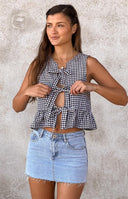 A close-up of a black and white gingham print sleeveless top featuring a deep V-neckline with multiple tie-front closures and a ruffled hem.