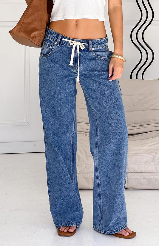Model wearing stylish wide-leg blue denim jeans with a drawstring waist, paired with a white crop top and brown accessories.