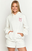 Brunette girl wearing a big oversized white hoodie with pink bow designs embroidered on the chest and sleeves. Girl is wearing white ruffled shorts.