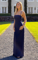Navy blue satin maxi dress with thin spaghetti straps, a square neckline, and a low back with criss-cross detailing. A light blue bracelet is worn.