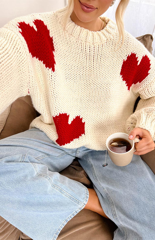 a blonde girl wears a white knit sweater with red hearts, denim jeans and holding a cup of tea