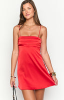 Model posing in a vibrant red satin mini dress suitable for homecoming. The dress features a straight, ruched neckline and thin spaghetti straps for a chic, formal look.