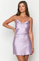 Model wearing purple satin mini dress in front of white curtains.