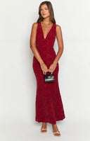 a blonde model wears a red maxi dress and gold bracelet holding a red rose, with red roses in the background on a table