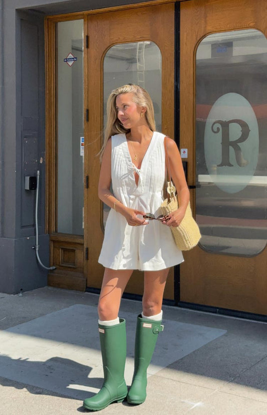 A woman stands outdoors in a sleeveless white playsuit featuring a V-neck with a tie at the bust and vertical pintuck detailing. She is wearing tall, matte green rain boots over white socks that peek out at the top.