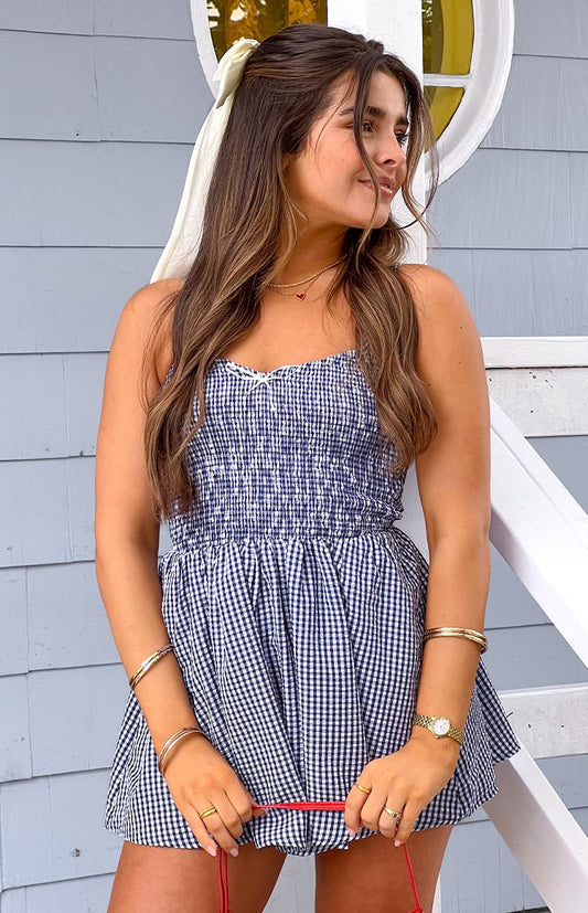 Model is wearing a navy and white gingham playsuit featuring a shirred bodice, thin shoulder straps, and a small white bow detail at the neckline. Her hair is accented with a cream bow.