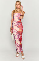Girl looking back, highlighting the side and back view of a strapless floral maxi dress in shades of purple and red, emphasising its continuous side ruching for a sculpted fit and the elegant sheer overlay of the skirt.