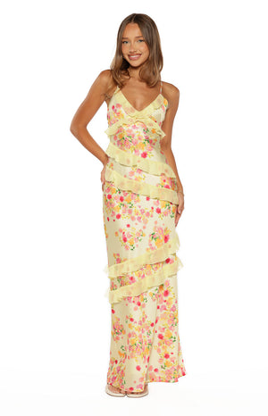 A full-length studio shot of our model wearing a pale yellow maxi dress with a vibrant pink orange and yellow floral print. This maxi dress features thin spaghetti straps a V-neckline and several tiers of sheer diagonal ruffles flowing down the body.