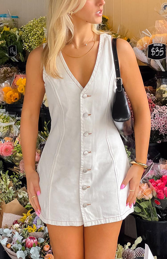 A versatile white wash denim mini dress, designed with a V-neckline and a full-length button-front closure, accented with contrasting stitching, suitable for a casual outing.
