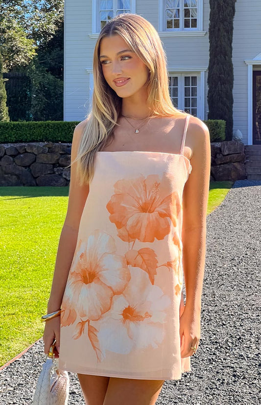 A light peach mini dress adorned with large, soft orange and white hibiscus floral prints, featuring a simple square neckline and delicate spaghetti straps, suitable for a casual summer event.