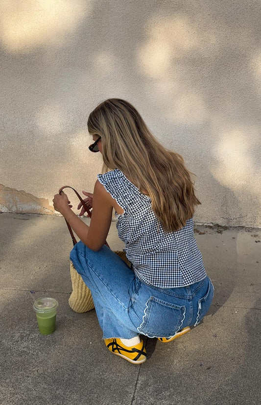  A woman crouches to show the back detail of her mid-wash jeans, which feature a white scalloped embroidered trim along the top of the back pockets. She is wearing a blue and white gingham top and yellow sneakers.