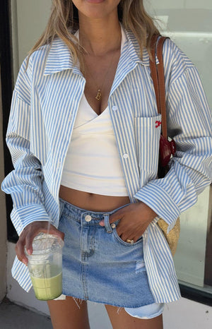 Model wearing an oversized blue and white striped button-up shirt, styled open over a white top and a denim mini skirt. The shirt has a small red embroidered detail on the chest pocket.
