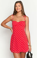 Model wearing a vibrant red mini dress with white polka dots, a sweetheart neckline, and thin tie-straps, accessorized with gold jewelry and a dark bag.