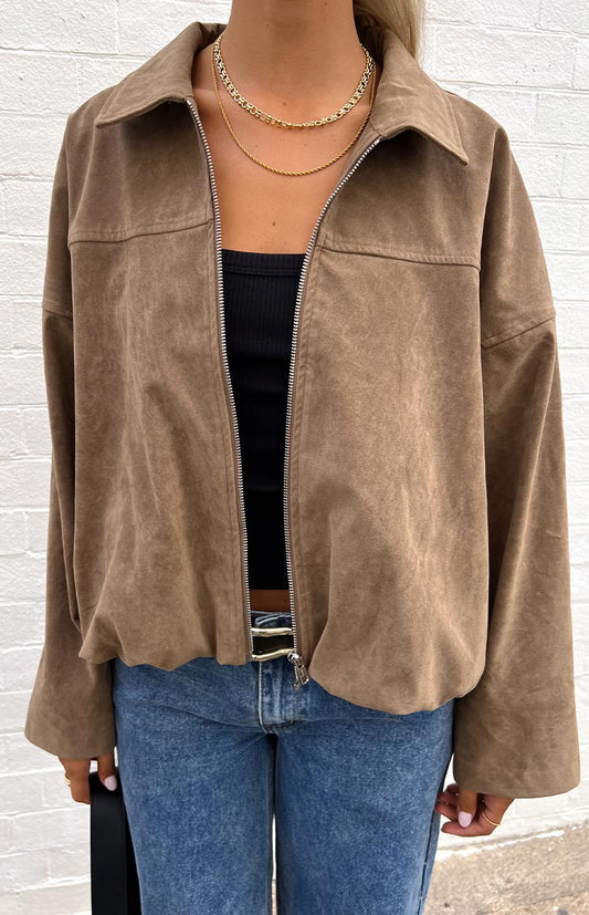 Close-up of a model wearing an open brown suede-like or corduroy jacket over a black ribbed top, with blue jeans and gold layered necklaces.