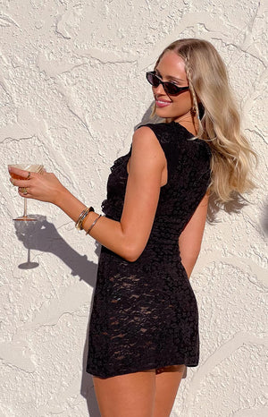 Girl in a black lace mini dress looks over her shoulder with a smile, holding a cocktail glass, showcasing the dress's texture and her gold bangles against a sunlit white wall.