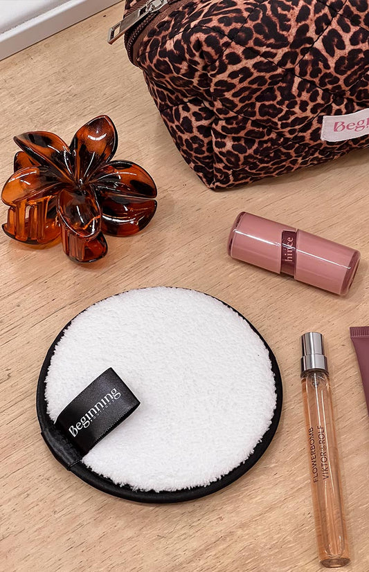 A round, white reusable makeup removing pad with a black border and a black 'Beginning' label, lying on a wooden surface with other beauty products.