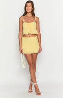 Brunette model wearing a yellow two piece set, flowy crop top and mini skirt with a gold necklace holding a drink.